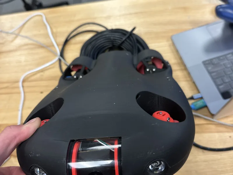 The Tryton hull in one hand. A 3D printed black body with red fittings,
                  open thruster nacelles and a clear acrylic tube holding the camera.
